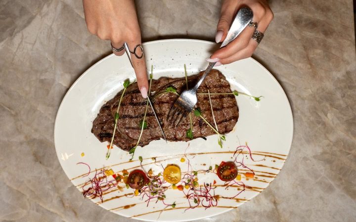 Une personne découpe une pièce de viande grillée sur une assiette décorée de légumes et de sauce