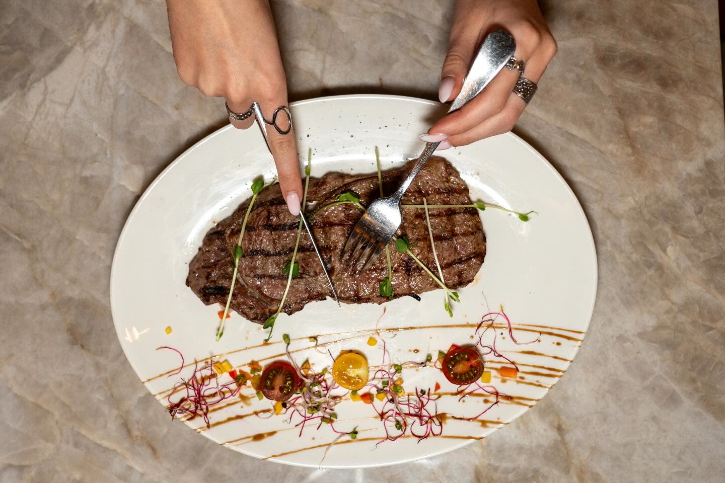 Une personne découpe une pièce de viande grillée sur une assiette décorée de légumes et de sauce